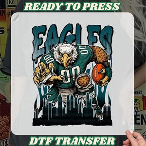 May include: White DTF transfer with a cartoon eagle in a football uniform, clutching a football. The word "EAGLES" is above the eagle, with the text "READY TO PRESS" and "DTF TRANSFER" also present.