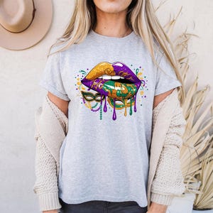 May include: A light gray t-shirt featuring a graphic of stylized lips in gold, purple, and green, with a Mardi Gras mask design. The lips have dripping paint effects and are decorated with colorful accents.