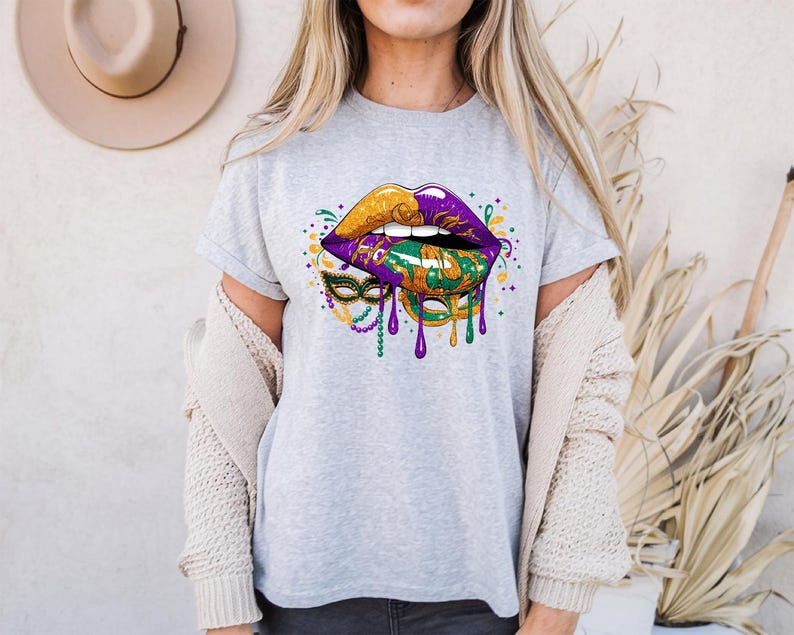 May include: A light gray t-shirt featuring a graphic of stylized lips in gold, purple, and green, with a Mardi Gras mask design. The lips have dripping paint effects and are decorated with colorful accents.