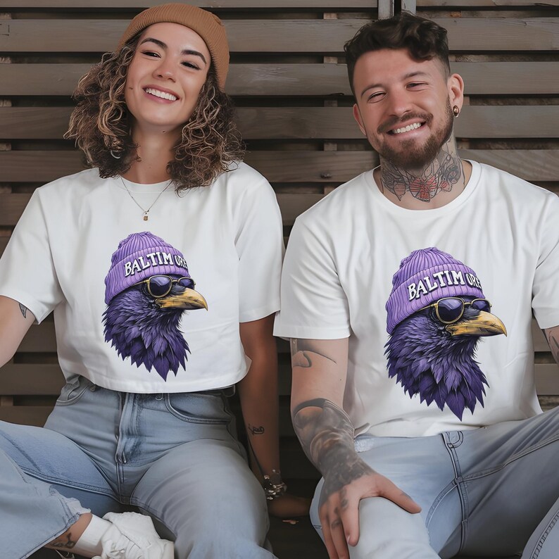 Pu&ograve; includere: T-shirt bianche con un disegno di corvo viola che indossa un berretto viola con la scritta "Baltim" e occhiali da sole. Il disegno del corvo &egrave; centrato sulla parte anteriore delle magliette. Le persone nell'immagine sorridono.