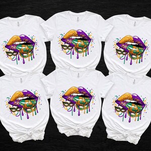 May include: Six white t-shirts with a colorful graphic of lips, a mask, and beads. The lips are gold and purple, with green and gold accents. The mask is green and gold, and the beads are purple and gold. The design has a dripping effect.