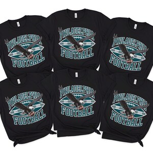 Może przedstawiać: Sześć czarnych koszulek z napisem "PHILADELPHIA FOOTBALL" w kolorze białym i morskim. Koszulki przedstawiają grafikę orła trzymającego piłkę nożną. Koszulki są z kr&oacute;tkim rękawem.