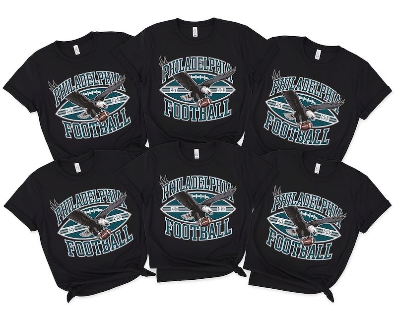 Może przedstawiać: Sześć czarnych koszulek z napisem "PHILADELPHIA FOOTBALL" w kolorze białym i morskim. Koszulki przedstawiają grafikę orła trzymającego piłkę nożną. Koszulki są z kr&oacute;tkim rękawem.