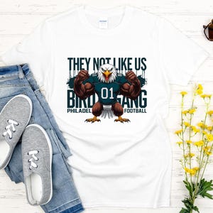 K&ouml;nnte beinhalten: Wei&szlig;es T-Shirt mit einer Grafik eines muskul&ouml;sen Adlers, der ein Football-Trikot mit der Nummer 01 tr&auml;gt. Auf dem Shirt steht "THEY NOT LIKE US BIRD GANG PHILADELPHIA FOOTBALL."