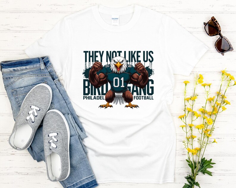 K&ouml;nnte beinhalten: Wei&szlig;es T-Shirt mit einer Grafik eines muskul&ouml;sen Adlers, der ein Football-Trikot mit der Nummer 01 tr&auml;gt. Auf dem Shirt steht "THEY NOT LIKE US BIRD GANG PHILADELPHIA FOOTBALL."