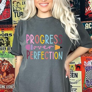 May include: Dark gray t-shirt with the words "PROGRESS over PERFECTION" in colorful letters and a pencil graphic. The shirt is a crew neck and has short sleeves. The background features vintage concert posters.