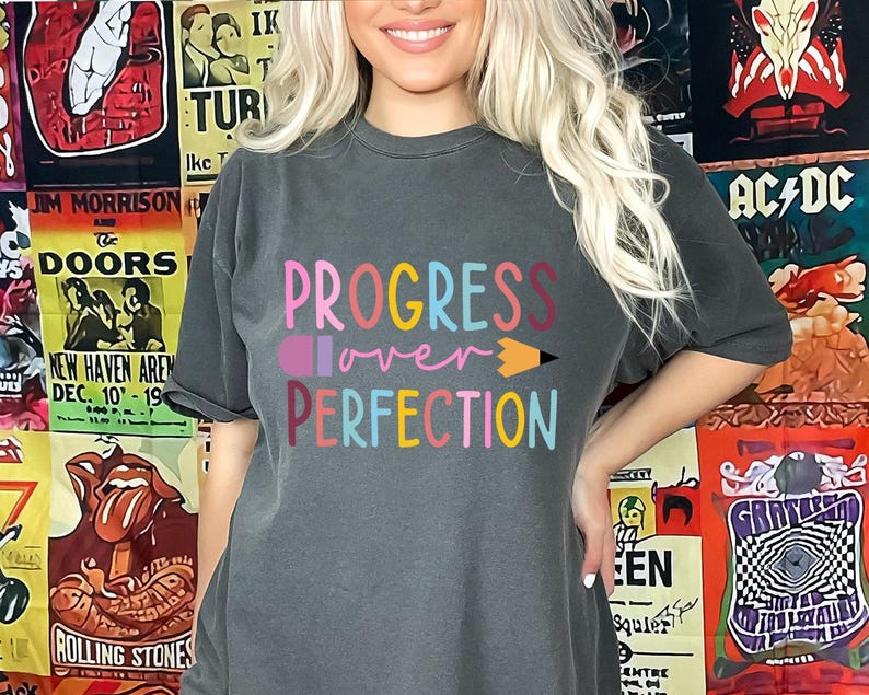 May include: Dark gray t-shirt with the words "PROGRESS over PERFECTION" in colorful letters and a pencil graphic. The shirt is a crew neck and has short sleeves. The background features vintage concert posters.