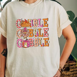 May include: Cream-colored t-shirt with the word "GOBBLE" in three lines. Each line features a different Thanksgiving-themed graphic: a pie, a turkey, and a pumpkin. The letters are in orange, pink, and white checkered patterns.