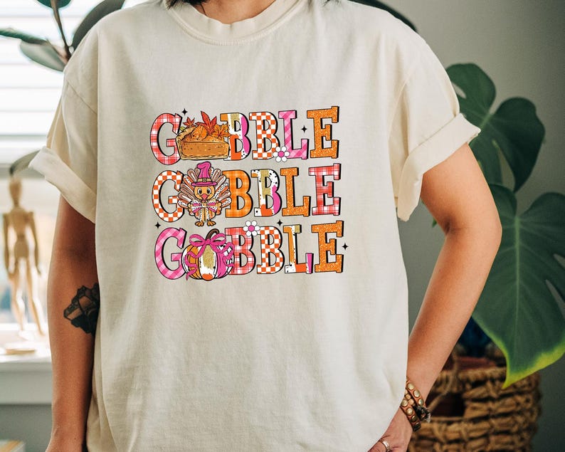May include: Cream-colored t-shirt with the word "GOBBLE" in three lines. Each line features a different Thanksgiving-themed graphic: a pie, a turkey, and a pumpkin. The letters are in orange, pink, and white checkered patterns.