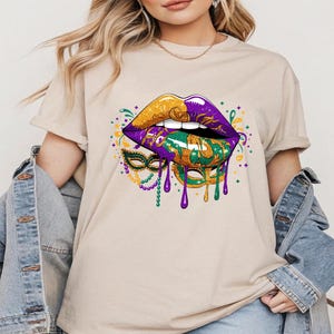 May include: Beige t-shirt featuring a vibrant graphic of stylized lips in gold, purple, and green, with a matching mask and dripping paint effect. The design is reminiscent of Mardi Gras, with a festive and colorful aesthetic.