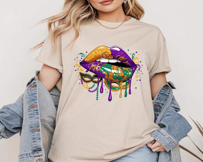 May include: Beige t-shirt featuring a vibrant graphic of stylized lips in gold, purple, and green, with a matching mask and dripping paint effect. The design is reminiscent of Mardi Gras, with a festive and colorful aesthetic.