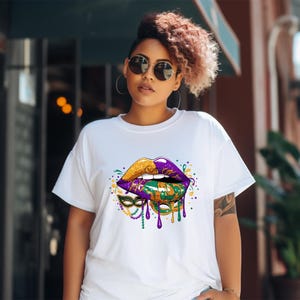 May include: White t-shirt featuring a vibrant graphic of stylized lips in gold, purple, and green, with a Mardi Gras mask design. The design includes dripping paint effects and colorful accents, creating a festive and eye-catching look.