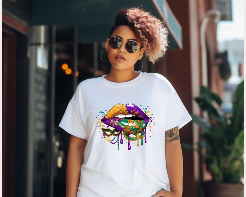 May include: White t-shirt featuring a vibrant graphic of stylized lips in gold, purple, and green, with a Mardi Gras mask design. The design includes dripping paint effects and colorful accents, creating a festive and eye-catching look.