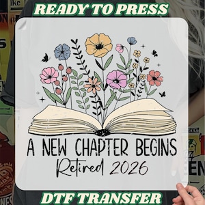 May include: A DTF transfer design with an open book illustration, adorned with vibrant flowers and butterflies. The text includes "A NEW CHAPTER BEGINS Retired 2026", "READY TO PRESS", and "DTF TRANSFER" in green.