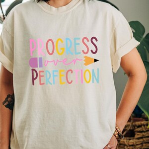 May include: Cream-colored t-shirt with the words "PROGRESS over PERFECTION" in colorful, block letters. A pencil graphic replaces the letter "I" in "PERFECTION". The shirt has rolled-up sleeves and a relaxed fit.