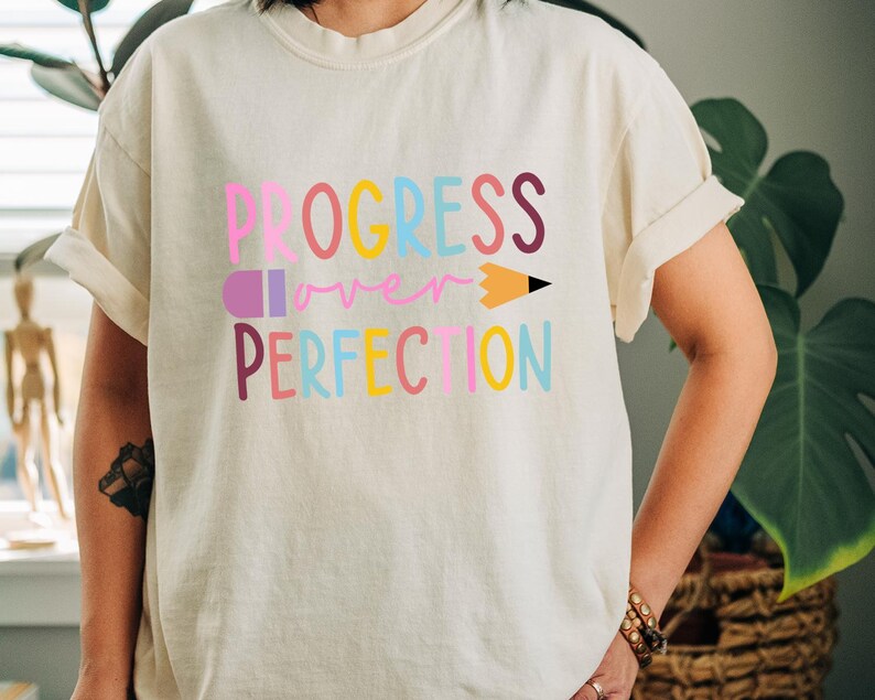 May include: Cream-colored t-shirt with the words "PROGRESS over PERFECTION" in colorful, block letters. A pencil graphic replaces the letter "I" in "PERFECTION". The shirt has rolled-up sleeves and a relaxed fit.