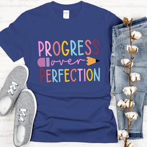 May include: A navy blue t-shirt with the words "PROGRESS over PERFECTION" in colorful letters and a pencil graphic. The shirt is paired with gray sneakers and blue jeans, with a sprig of cotton blossoms.