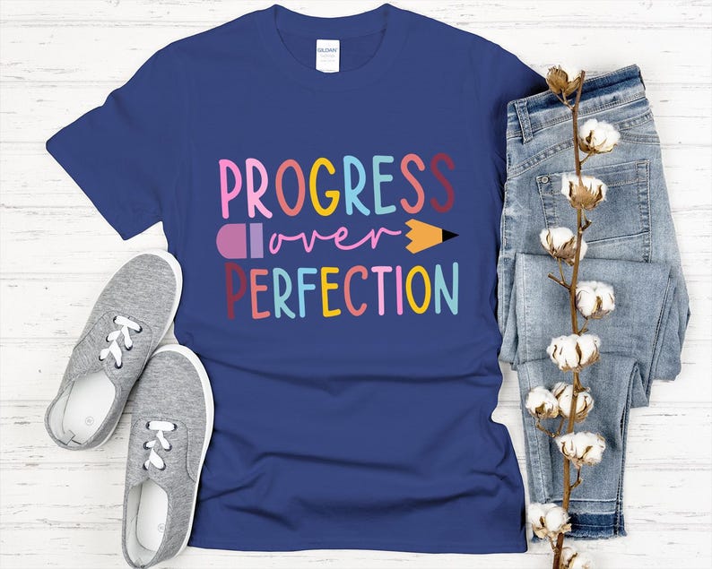 May include: A navy blue t-shirt with the words "PROGRESS over PERFECTION" in colorful letters and a pencil graphic. The shirt is paired with gray sneakers and blue jeans, with a sprig of cotton blossoms.
