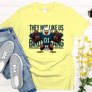 K&ouml;nnte beinhalten: Ein hellgelbes T-Shirt mit einem muskul&ouml;sen Adler, der ein Football-Trikot mit der Nummer 01 tr&auml;gt. Auf dem Shirt steht "THEY NOT LIKE US BIRD GANG PHILADELPHIA FOOTBALL". Das Shirt wird mit blauen Jeans und grauen Turnschuhen kombiniert.