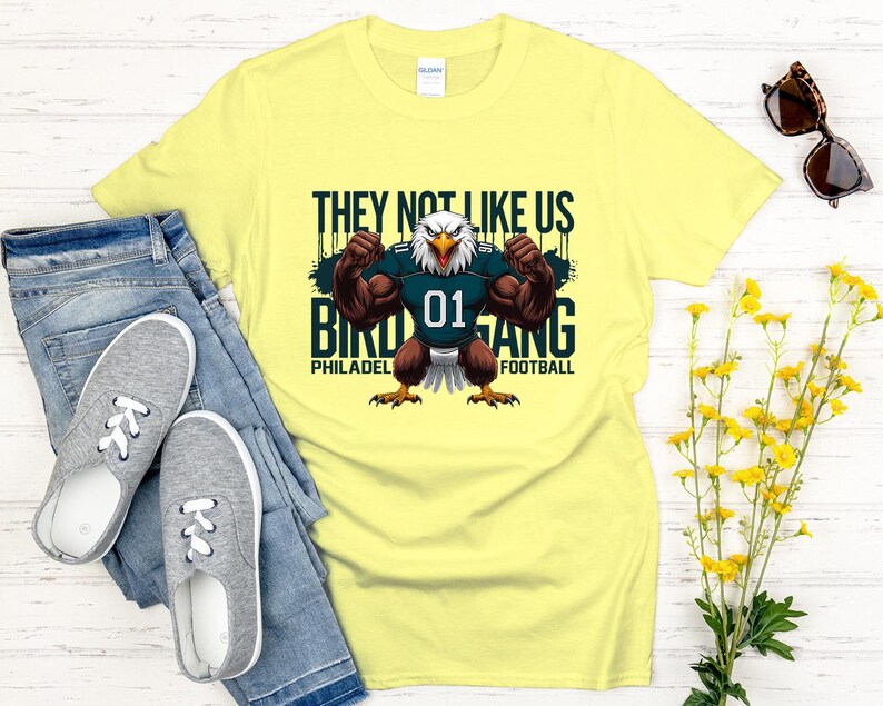 K&ouml;nnte beinhalten: Ein hellgelbes T-Shirt mit einem muskul&ouml;sen Adler, der ein Football-Trikot mit der Nummer 01 tr&auml;gt. Auf dem Shirt steht "THEY NOT LIKE US BIRD GANG PHILADELPHIA FOOTBALL". Das Shirt wird mit blauen Jeans und grauen Turnschuhen kombiniert.