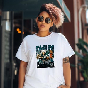 May include: White t-shirt with a graphic of an eagle in a football uniform and the word "EAGLES". The eagle is holding a football. The design uses blue, green, and brown colours.