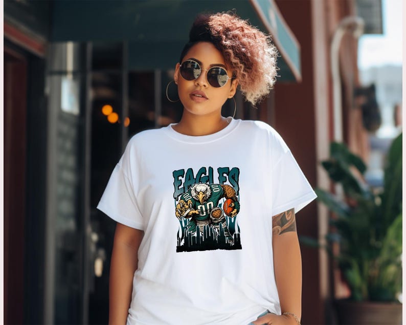 May include: White t-shirt with a graphic of an eagle in a football uniform and the word "EAGLES". The eagle is holding a football. The design uses blue, green, and brown colours.