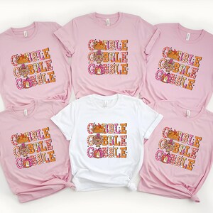May include: A collection of pink and white t-shirts with the text "GOBBLE GOBBLE GOBBLE" in orange, pink, and white. The graphic includes a turkey, a pumpkin, and fall leaves. The shirts are short-sleeved.