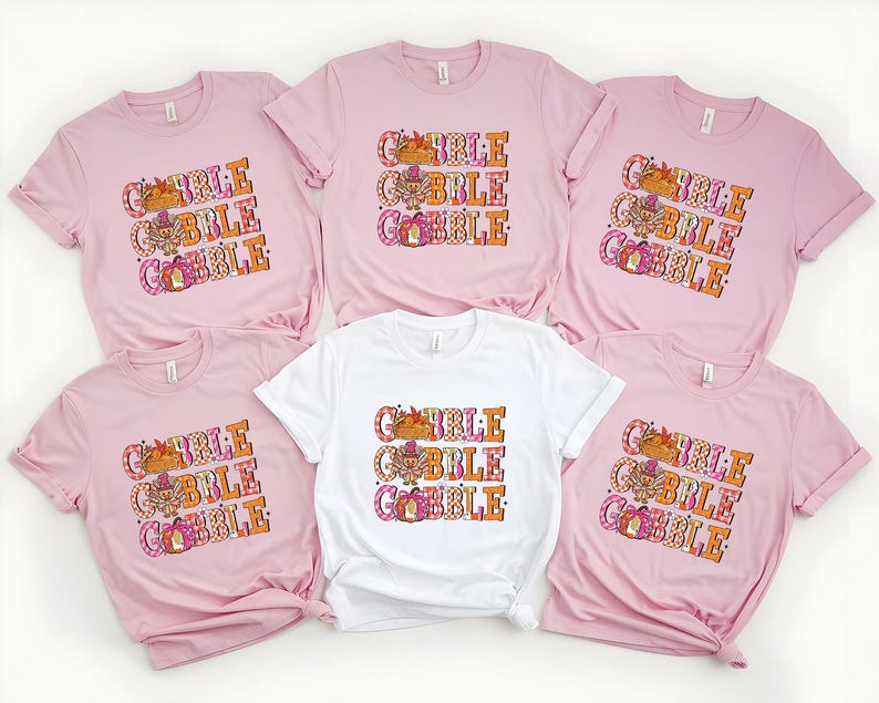 May include: A collection of pink and white t-shirts with the text "GOBBLE GOBBLE GOBBLE" in orange, pink, and white. The graphic includes a turkey, a pumpkin, and fall leaves. The shirts are short-sleeved.
