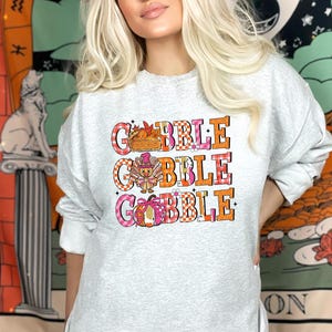 May include: A light gray sweatshirt with the words "GOBBLE GOBBLE GOBBLE" in orange and pink block letters. The letters are layered over a plaid pattern. The design includes a basket of food, a turkey, and a pumpkin.