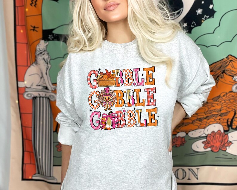 May include: A light gray sweatshirt with the words "GOBBLE GOBBLE GOBBLE" in orange and pink block letters. The letters are layered over a plaid pattern. The design includes a basket of food, a turkey, and a pumpkin.