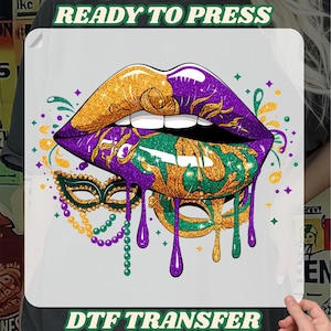 May include: DTF transfer featuring a vibrant design of lips in gold, purple, and green, with a dripping effect. A masquerade mask and beaded necklace are also included in the design. The text "READY TO PRESS" and "DTF TRANSFER" are displayed.