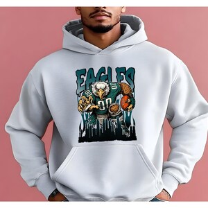 May include: White hoodie with a graphic of an eagle in a football uniform, holding a football. The word "EAGLES" is printed above the eagle in teal letters. The design includes a cityscape silhouette at the bottom.