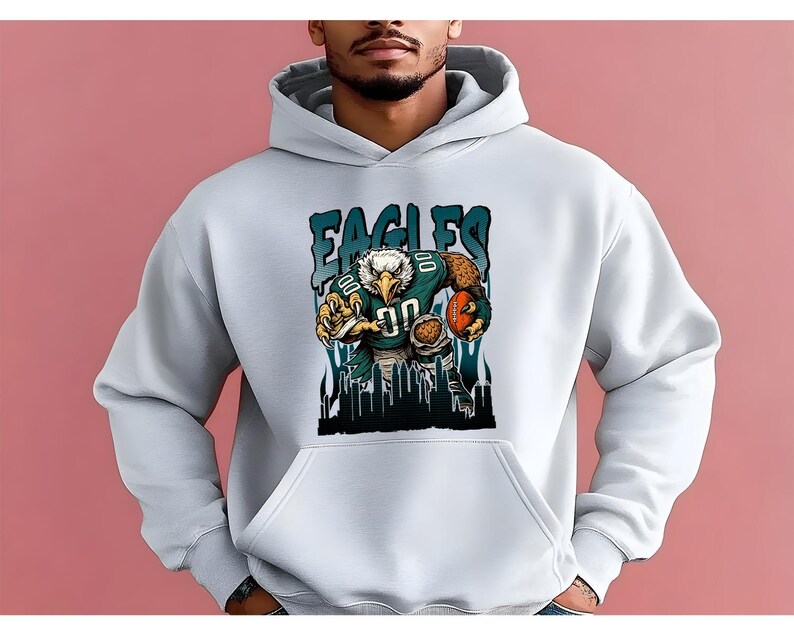 May include: White hoodie with a graphic of an eagle in a football uniform, holding a football. The word "EAGLES" is printed above the eagle in teal letters. The design includes a cityscape silhouette at the bottom.