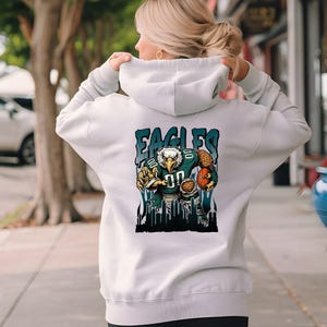 May include: A pale grey hoodie with a graphic of an eagle in a football uniform, with the text "EAGLES". The eagle is holding a football. The hoodie has a drawstring hood.