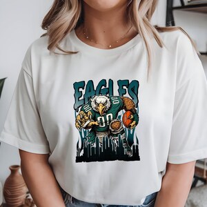 May include: White t-shirt with a cartoon eagle in a football uniform, holding a football. The word "EAGLES" is printed above the eagle in teal and black. The eagle is wearing the number "00".