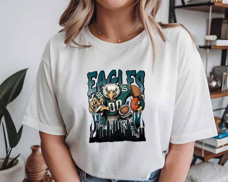 May include: White t-shirt with a cartoon eagle in a football uniform, holding a football. The word "EAGLES" is printed above the eagle in teal and black. The eagle is wearing the number "00".