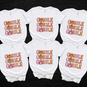 May include: Six white t-shirts with the text "GOBBLE GOBBLE GOBBLE" in orange, pink, and brown. The design includes a turkey, pumpkins, and fall leaves, perfect for Thanksgiving. The shirts are short-sleeved.