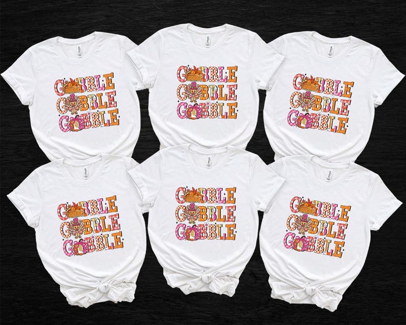 May include: Six white t-shirts with the text "GOBBLE GOBBLE GOBBLE" in orange, pink, and brown. The design includes a turkey, pumpkins, and fall leaves, perfect for Thanksgiving. The shirts are short-sleeved.