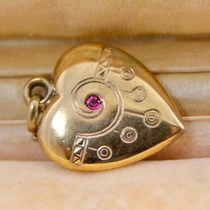 Può includere: Ciondolo a forma di cuore color oro con una piccola gemma rosa rotonda al centro. Il ciondolo presenta un design inciso con forme geometriche e cerchi. Un piccolo anello è attaccato per una catena.