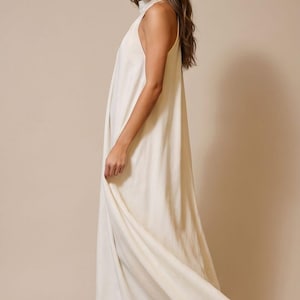 May include: A floor-length, off-white halter dress. The dress has a high neckline and a flowing, loose fit. The fabric appears to be a lightweight material. The model is wearing neutral-colored sandals.