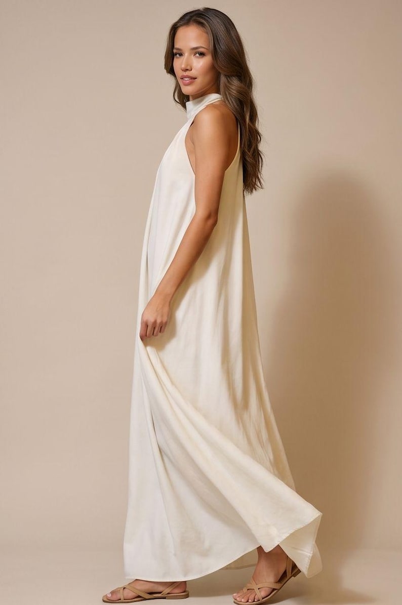 May include: A floor-length, off-white halter dress. The dress has a high neckline and a flowing, loose fit. The fabric appears to be a lightweight material. The model is wearing neutral-colored sandals.