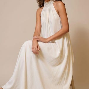 May include: A cream-colored halter neck maxi dress. The dress features a high neckline and a flowing skirt. The model is seated on a wooden stool, wearing sandals. The dress is made of a soft, flowing fabric.