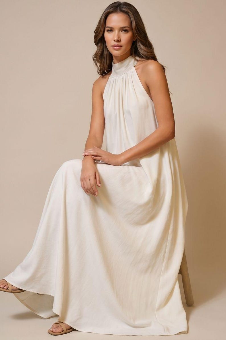 May include: A cream-colored halter neck maxi dress. The dress features a high neckline and a flowing skirt. The model is seated on a wooden stool, wearing sandals. The dress is made of a soft, flowing fabric.