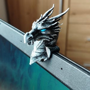 May include: A detailed, silver and black dragon head webcam cover with bright blue eyes. The dragon is perched on the edge of a laptop, adding a unique and decorative touch. The dragon's scales and horns are intricately designed.