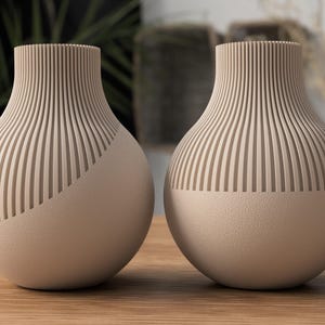 Könnte beinhalten: Zwei beigefarbene Keramikvasen mit einem gerippten Design. Die Vasen haben eine glatte, strukturierte Oberfläche auf der unteren Hälfte.