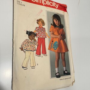 May include: Simplicity sewing pattern number 7604 for a toddler dress and pants. The pattern includes instructions for making an applique. The pattern shows two girls wearing the dresses and pants. One girl is wearing a red and white floral print dress and red pants. The other girl is wearing a yellow dress with a paint palette applique.