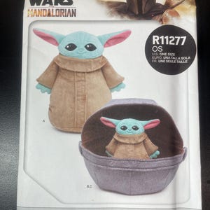 May include: A Simplicity home sewing pattern featuring a plush Grogu and soft space pram. The pattern includes images of the Grogu plush toy in a tan coat with large ears and a pram. The Star Wars Mandalorian logo is at the top.