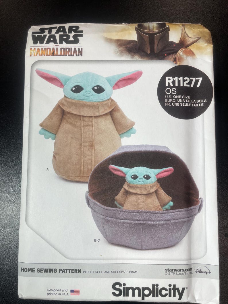 May include: A Simplicity home sewing pattern featuring a plush Grogu and soft space pram. The pattern includes images of the Grogu plush toy in a tan coat with large ears and a pram. The Star Wars Mandalorian logo is at the top.