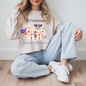 Pode incluir: Camisola bege com o texto "A Programme" e um gráfico do espaço de trabalho de um programador. O gráfico inclui um laptop, uma secretária e símbolos de código. A frase "Eat. Sleep. Code. Repeat." está impressa abaixo.