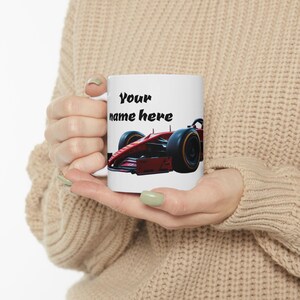 Puede incluir: Taza de cerámica blanca con un gráfico de coche de carreras de Fórmula 1 rojo y el texto "Your name here".
